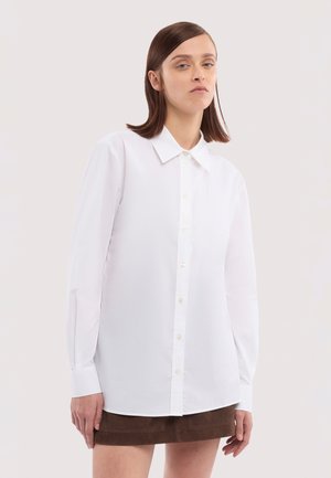 Vrouw met stijlhaar, bruin haar, die een witte blouse met lange mouwen en een bruine rok draagt, staand tegen een effen lichte achtergrond.