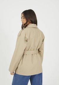 Beżowa kurtka wierzchnia z paskiem, z klasycznym kołnierzem, wykonana z gładkiego, lekkiego materiału, prezentowana z tyłu. Posiada luźne rękawy.