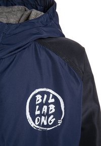 Granatowa kurtka z szarym wykończeniem, z okrągłym białym logo na klatce piersiowej. Materiał wydaje się gładki z delikatnie błyszczącą fakturą.