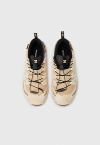 Des chaussures de sport beiges avec une tige en tissu texturé, un col en mesh noir, des lacets élastiques et une semelle en caoutchouc présentant un motif de sculpture.
