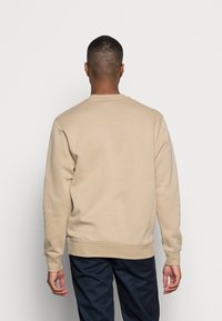 Sudadera de cuello redondo beige hecha de material suave, con puños y dobladillo acanalados. Llevada con pantalones azul marino, vista desde la parte de atrás.