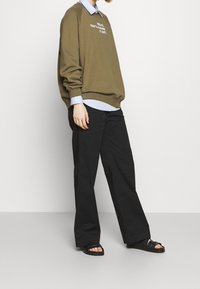 Felpa verde oliva con testo bianco, indossata sopra una camicia colletto blu chiaro, abbinata a pantaloni larghi neri e ciabatte nere.