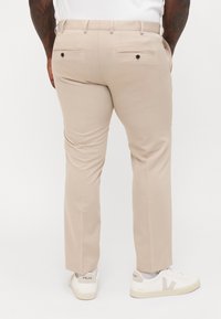 Beige tailored trousers with a smooth texture, two back pockets with black buttons, worn with white sneakers featuring a gray V logo.