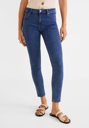 Femme portant un jean skinny bleu foncé, un haut beige sans manches et des sandales beiges à deux brides, debout devant un fond blanc.