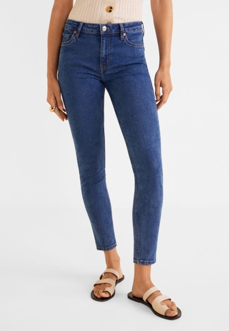 Femme portant un jean skinny bleu foncé, un haut beige sans manches et des sandales beiges à deux brides, debout devant un fond blanc.