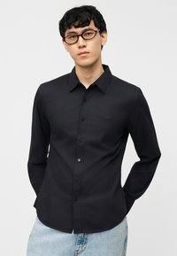 Άνδρας με μαύρα σγουρά μαλλιά, φορώντας μαύρο πουκάμισο με κουμπιά, γυαλιά και ανοιχτόχρωμο τζιν, στέκεται με τα χέρια πίσω από την πλάτη σε απλό φόντο.