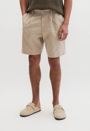 Mann trägt beige Shorts, braunes Strickoberteil und beige Slipper und steht vor einem einfarbigen Hintergrund.