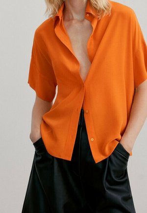 Person wearing an unbuttoned bright orange short-sleeve shirt with hands in black wide-leg pants pockets against a plain background.