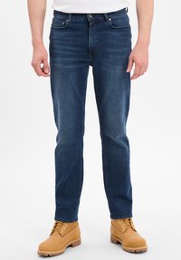 Ciemnoniebieskie dżinsy o slim fit z delikatnym przetarciem, pięcioma kieszeniami i zapięciem na guzik, noszone z beżowymi trzewikami na sznurówki.