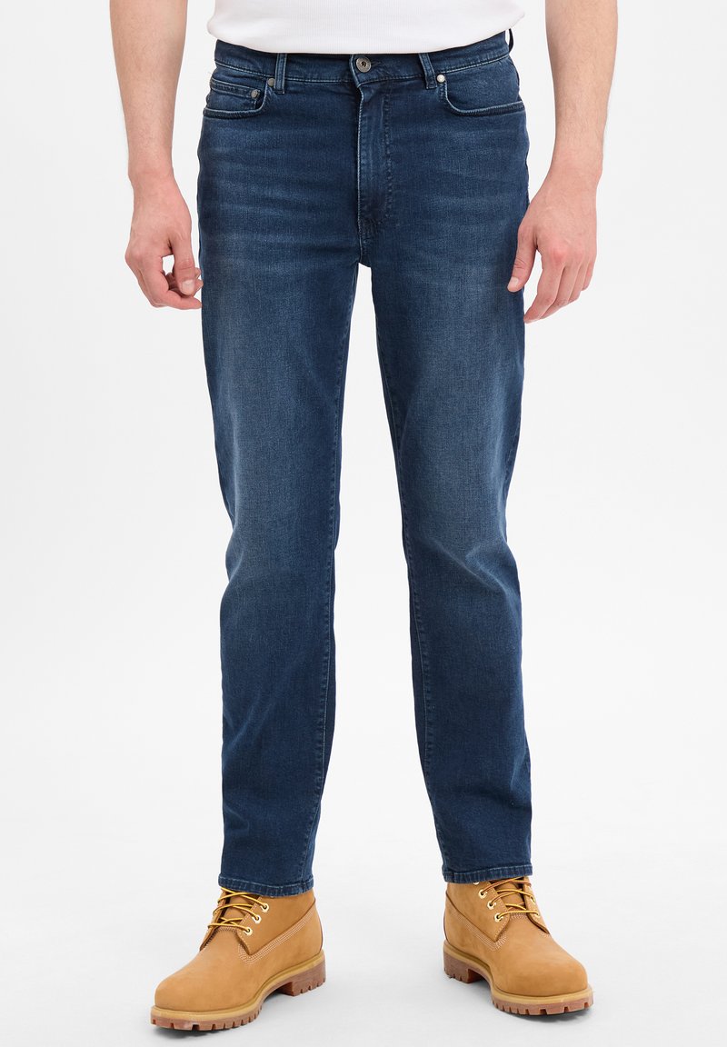 Ciemnoniebieskie dżinsy o slim fit z delikatnym przetarciem, pięcioma kieszeniami i zapięciem na guzik, noszone z beżowymi trzewikami na sznurówki.