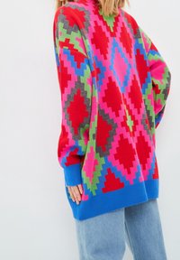 Kolorowy, oversize'owy sweter prezentujący geometryczny wzór w kolorach czerwonym, różowym, zielonym i niebieskim. Wykonany z miękkiego materiału dzianinowego z prążkowanymi mankietami.