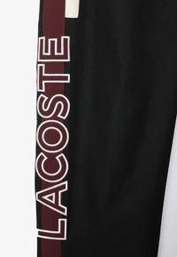 Svarta träningsbyxor med en vinröd sidrand och fet vit "LACOSTE"-text på vänster ben, tillverkade av ett strukturerat material.
