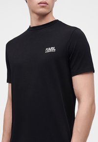 Άνδρας που φοράει μαύρο κοντομάνικο T-shirt με λευκό λογότυπο "Karl Lagerfeld" τυπωμένο στο αριστερό στήθος.