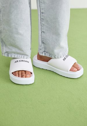 Valged sandaalid avatud varvastega kujundusega, mille rihmal on musta teksti "AIR JORDAN" ja paks valge tald.