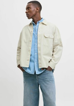 Man wearing light beige button-up jacket over light blue denim shirt and jeans, hands in pockets, looking to the left against plain background.