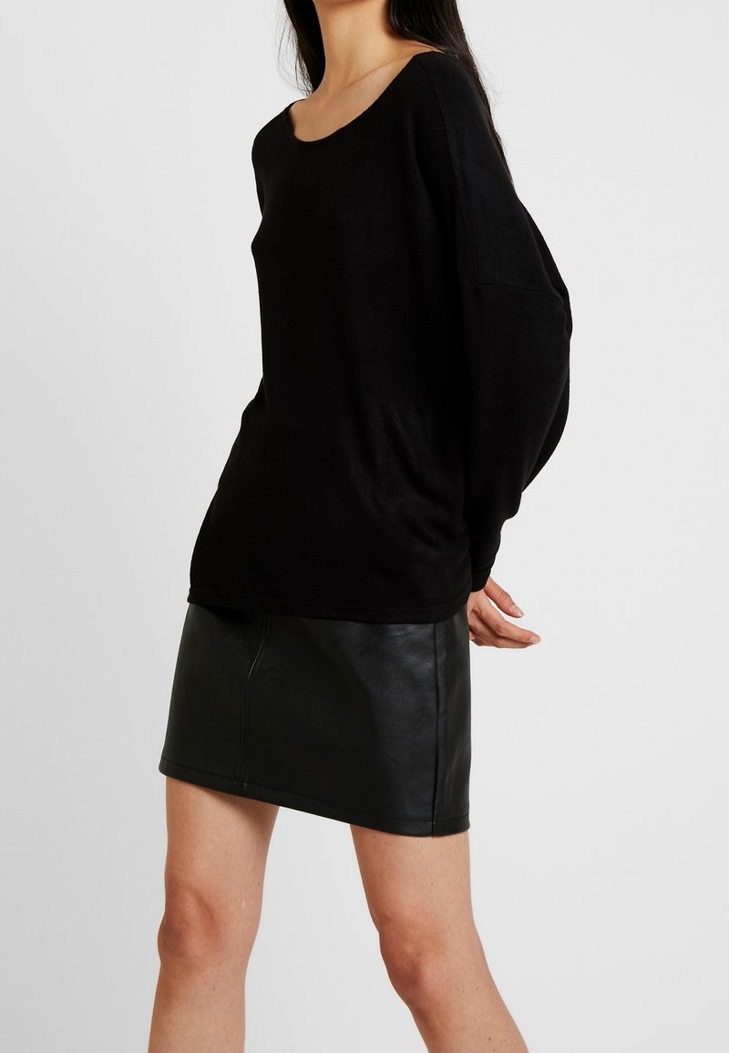 Femme portant un haut noir à manches longues et ample, rentré dans une mini-jupe en cuir noir, posant devant un fond clair uni.