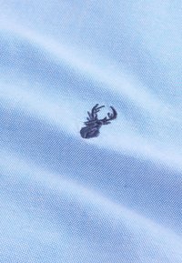 Lichtblauw stof met een subtiel gestippeld patroon. Voorzien van een geborduurd donkerblauw krabmotief, centraal op de stof geplaatst.