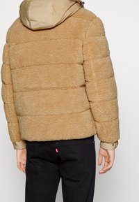 Beige teddyjas met textuurstof, ritsluiting en een capuchon. Zwarte broek met een zichtbaar logo-label op het achterzak.