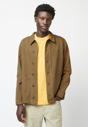 Brown canvas jacket with a button-up front, large pockets, and a classic collar, worn over a yellow shirt and light-colored pants.