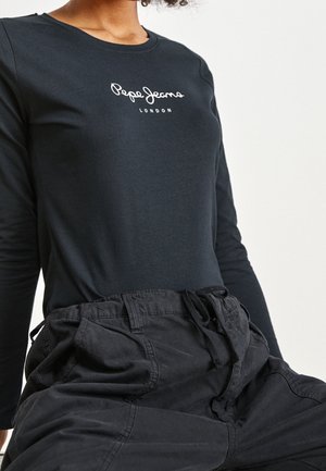 Personne portant un t-shirt noir à manches longues Pepe Jeans London et un pantalon noir ample avec une taille à cordon, assise devant un fond blanc.