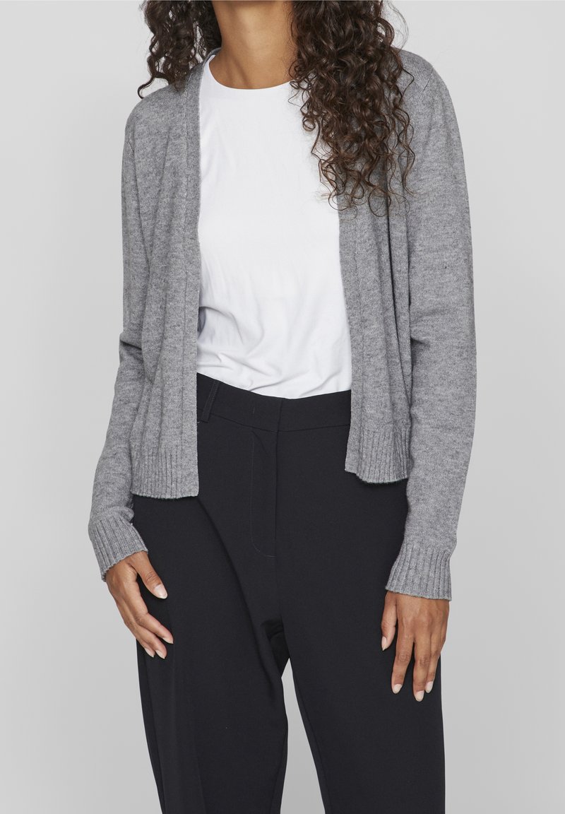 Grå stickad cardigan med ribbad fåll och manschetter, öppen framdesign, buren över en vit skjorta och svarta byxor.