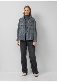 Light blue corduroy jacket with two chest pockets, button closure, worn over a gray turtleneck, paired with dark wide-leg jeans and cream sneakers.