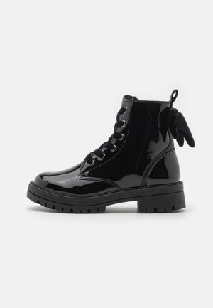 Bottine en cuir verni noir brillant avec semelle épaisse, lacets noirs et un nœud noir décoratif à l'arrière.
