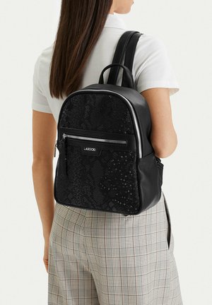 Žena oděná do bílé krátkosvré košile a kostkovaných kalhot, zezadu, s černým texturovaným batohem Lasocki.