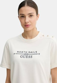 Jeune femme aux cheveux tirés en arrière portant un t-shirt blanc avec le texte "NORTH SAILS GUESS" et des détails de boutons sur l'épaule.