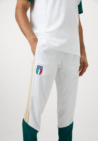 Άτομο που φορά λευκά ιταλικά ποδοσφαιρικά παντελόνια φόρμας με χρυσές ρίγες και πράσινα πάνελ, με τα χέρια στις τσέπες, μπροστά από απλή φόντο.