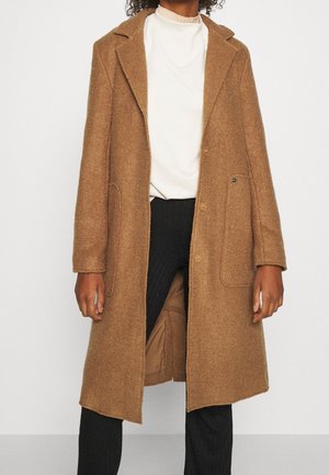 Femme portant un manteau en laine marron mi-long sur un haut blanc et un pantalon noir, debout devant un fond clair uni.