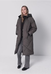 Long manteau matelassé vert olive avec capuche, présentant des coutures en forme de losange, une fermeture éclair et des poches latérales. Porté sur un pull noir et un jean gris.