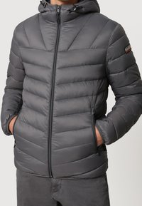 Man wearing a gray padded jacket with a hood, front zipper, side zippered pockets, and a small logo patch on the upper left sleeve.