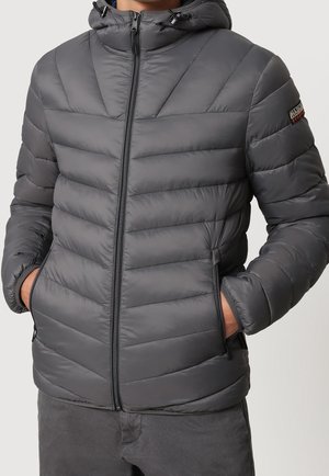 Man wearing a gray padded jacket with a hood, front zipper, side zippered pockets, and a small logo patch on the upper left sleeve.