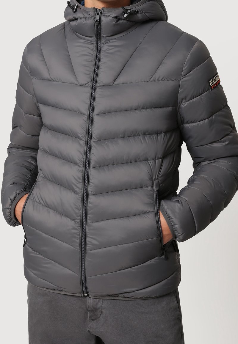 Man wearing a gray padded jacket with a hood, front zipper, side zippered pockets, and a small logo patch on the upper left sleeve.