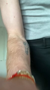 Forearm with light hair and skull tattoo, wearing a red and gold bracelet, next to a person in a gray shirt and black pants.