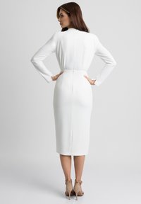Biała midi sukienka z długimi rękawami i dopasowanym stanem, charakteryzująca się gładką teksturą i zamek błyskawiczny z tyłu. Noszona z cielistymi szpilkami.