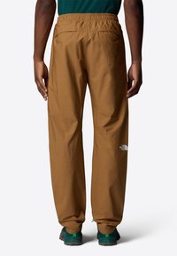Pantalons d'extérieur marron avec une taille élastique, présentant deux poches arrière et un logo blanc sur la jambe gauche. Le matériau semble léger.