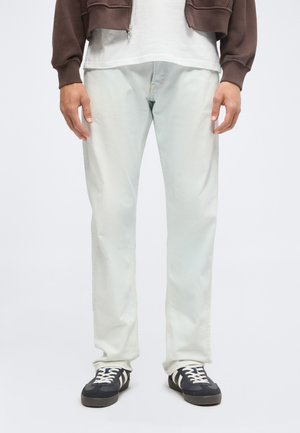 Homme portant un pantalon gris clair, des baskets noires et blanches, une chemise blanche et une veste marron à fermeture éclair, debout sur un fond uni.