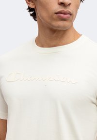 Νέος άνδρας με off-white μπλουζάκι Champion, δείχνοντας το στήθος και το κάτω μέρος του προσώπου σε απλό φόντο.
