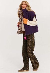 Fioletowa kamizelka z poduszkami, wykończona podszewką w leopardzie wzory, noszona na kremowym swetrze. Połączona z brązowymi spodniami w leopardzie wzory oraz brązowymi trampkami w paski.