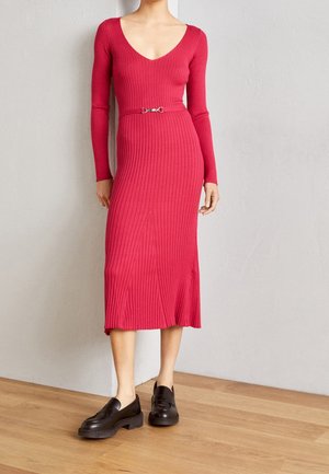 Kobieta ubrana w czerwoną, prążkowaną sukienkę midi z długim rękawem, z małym paskiem i czarnymi mokasynami, stojąca na drewnianej podłodze na tle jasnej ściany.