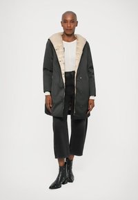 Parka imbottita con cappuccio beige, tessuto esterno verde scuro, zip frontale e chiusure a bottoni. Presenta tasche laterali e una vestibilità comoda.