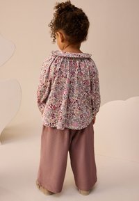 Virágmintás blúz fodros gallérral, széles, bordázott levendulaszínű nadrággal párosítva. Puha anyag, visszafogott színek és laza szabás.