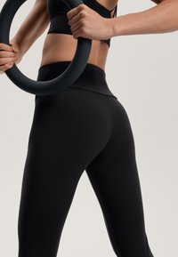 Czarne, dopasowane legginsy z wysokim stanem, zestawione z okrągłym czarnym akcesorium fitness trzymanym przez osobę. Gładka tekstura, minimalistyczny design.