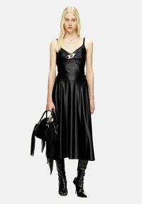 Abito in satin nero con corpetto aderente e gonna svasata, caratterizzato da un logo metallico sul petto. Abbinato a una borsa nera e stivali al ginocchio lucidi.