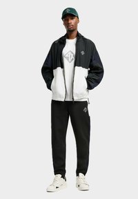 Giacca a vento bianca e nera con cappuccio, logo sul petto, abbinata a pantaloni neri e sneakers bianche. Un cappellino verde completa l'outfit.