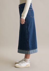 Długa spódnica dżinsowa w ciemnym niebieskim kolorze z jasnoniebieskim wykończeniem na dole, przednimi kieszeniami i dopasowaną talią. Zestawiona z białymi trampkami i beżowymi skarpetkami.