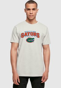 Šviesiai pilka medvilninė marškinė su mėlynu ir oranžiniu tekstu „Floridos universiteto Gators Gainesville, FL“, po kuriuo yra aligatoriaus grafika.