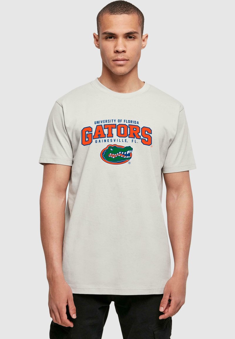 Šviesiai pilka medvilninė marškinė su mėlynu ir oranžiniu tekstu „Floridos universiteto Gators Gainesville, FL“, po kuriuo yra aligatoriaus grafika.
